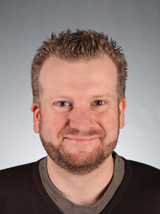 Tobias Kiessling mit kurzem Haar, Bart und dunklem Pullover, lächelnd.